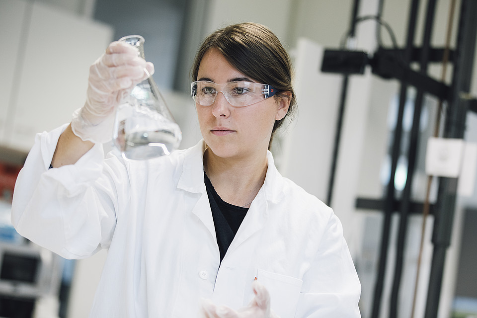 Studierende mit Schutzbrille, weißem Kittel und Handschuhen hält im Labor ein Reagenzglas hoch und betrachtet dessen Inhalt.