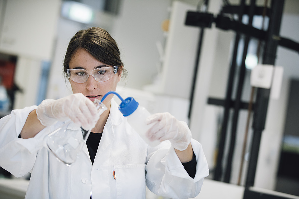 Studierende im weißen Kittel mit Schutzbrille arbeitet im Labor konzentriert mit einem Reagenzglas.